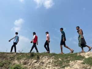 Five men walking on a grassy hill against a bright blue sky, showcasing casual outdoor fashion under a sunny climate.